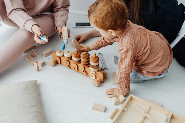 A young child engaged with a wooden toy train in a warm indoor setting, surrounded by adults.